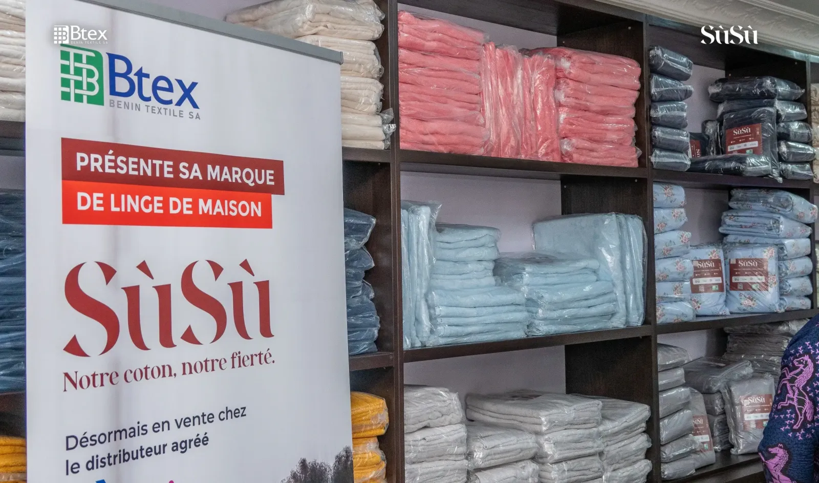Rayons de draps et de serviettes Sùsù dans la boutique, à côté d'une affiche Bénin Textile